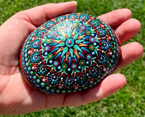 A hand holding out a rock painted with a blue and green mandala